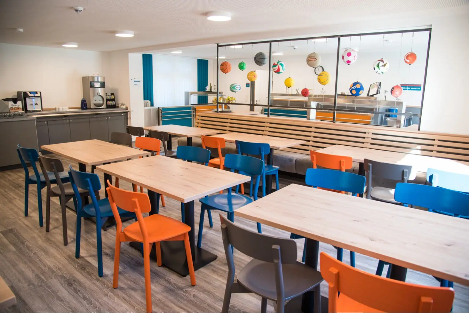 Foto, die die Inneneinrichtung des Speisaals der Sport Jugendherberge in Rheine, Holzstühle Mattis in bunt orange, blau und graubeige, Säulentische, helle HPL Tischplatten