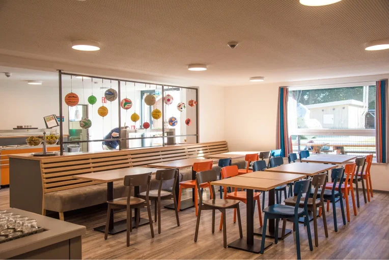 Foto, die die Inneneinrichtung des Speisaals der Sport Jugendherberge in Rheine, Holzstühle Mattis in bunt orange, blau und graubeige, Säulentische, helle HPL Tischplatten, Lattenbank mit Sitzpolster