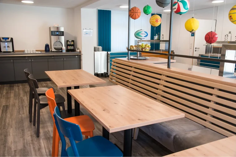 Foto, die die Inneneinrichtung des Speisaals der Sport Jugendherberge in Rheine, Holzstühle Mattis in bunt orange, blau und graubeige, Säulentische, helle HPL Tischplatten, Lattenbank mit Sitzpolster