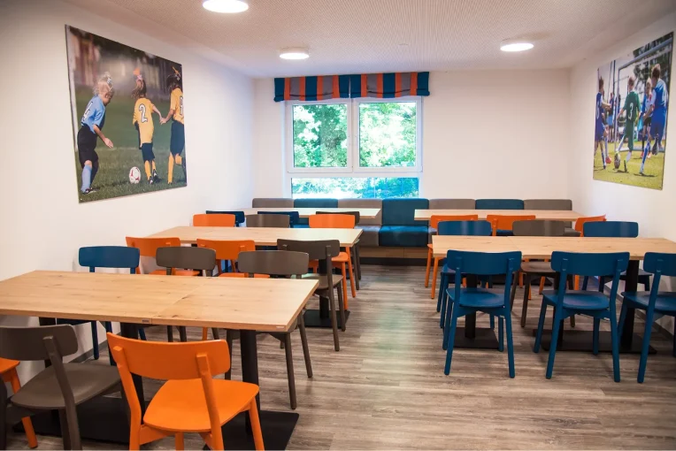 Foto, die die Inneneinrichtung des Speisaals der Sport Jugendherberge in Rheine, Holzstühle Mattis in bunt orange, blau und graubeige, Säulentische, helle HPL Tischplatten