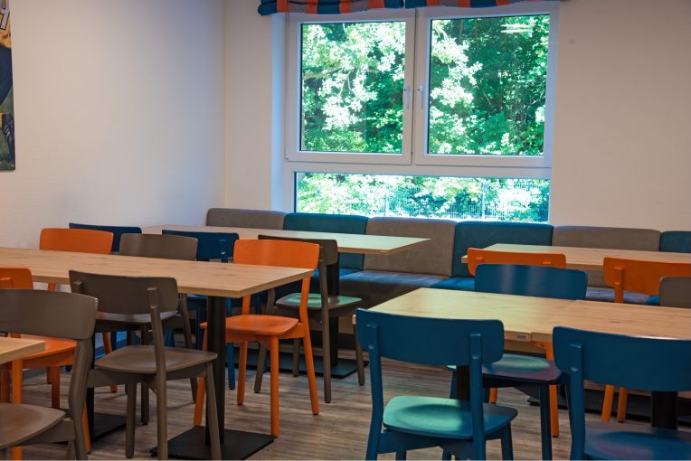 Foto, die die Inneneinrichtung des Speisaals der Sport Jugendherberge in Rheine, Holzstühle Mattis in bunt orange, blau und graubeige, Säulentische, helle HPL Tischplatten, Polsterbank im Hintergrund