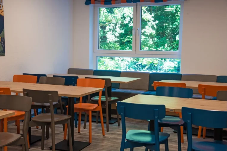 Foto, die die Inneneinrichtung des Speisaals der Sport Jugendherberge in Rheine, Holzstühle Mattis in bunt orange, blau und graubeige, Säulentische, helle HPL Tischplatten, Polsterbank im Hintergrund