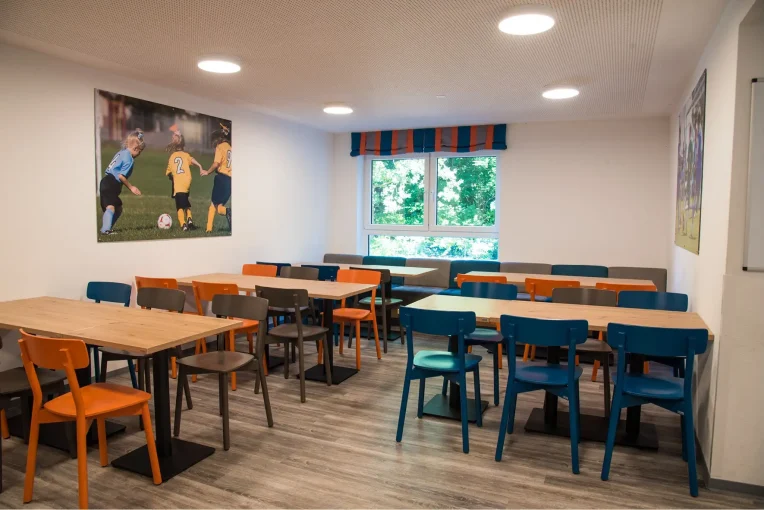 Foto, die die Inneneinrichtung des Speisaals der Sport Jugendherberge in Rheine, Holzstühle Mattis in bunt orange, blau und graubeige, Säulentische, helle HPL Tischplatten, Polsterbank im Hintergrund