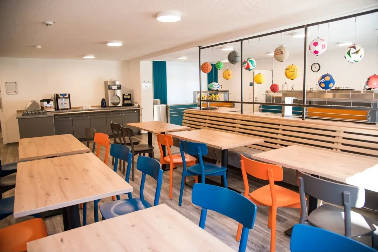 Foto, die die Inneneinrichtung des Speisaals der Sport Jugendherberge in Rheine, Holzstühle Mattis in bunt orange, blau und graubeige, Säulentische, helle HPL Tischplatten
