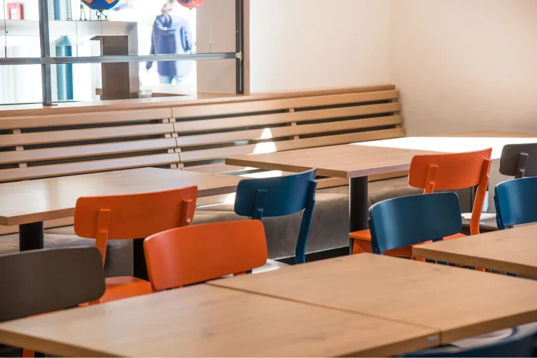 Foto, die die Inneneinrichtung des Speisaals der Sport Jugendherberge in Rheine, Holzstühle Mattis in bunt orange, blau und graubeige, Säulentische, helle HPL Tischplatten, Lattenbank