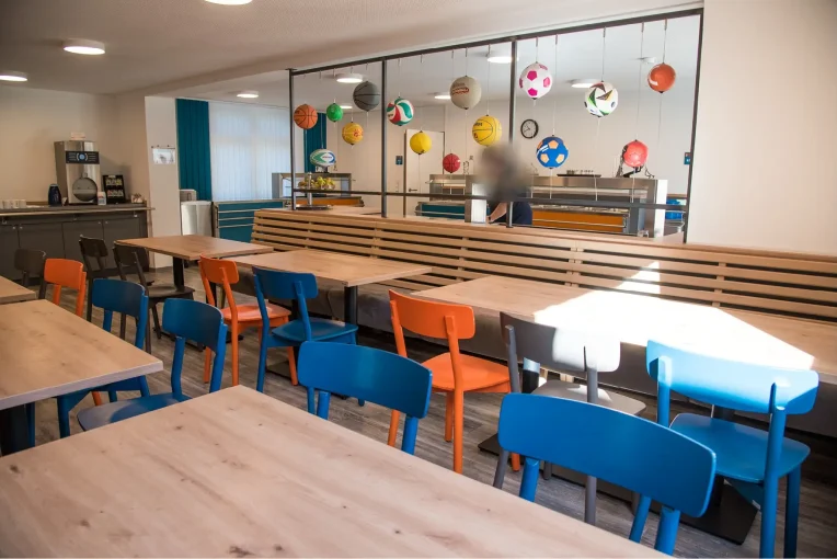 Foto, die die Inneneinrichtung des Speisaals der Sport Jugendherberge in Rheine, Holzstühle Mattis in bunt orange, blau und graubeige, Säulentische, helle HPL Tischplatten, Lattenbank mit Sitzpolster